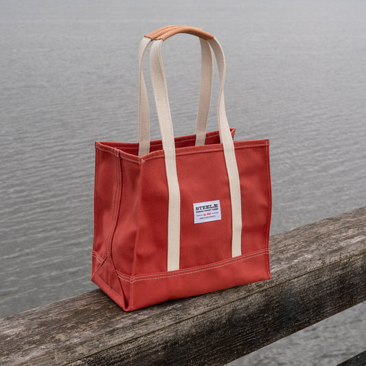 Red tote bag with beige handles on a wooden railing by a body of water