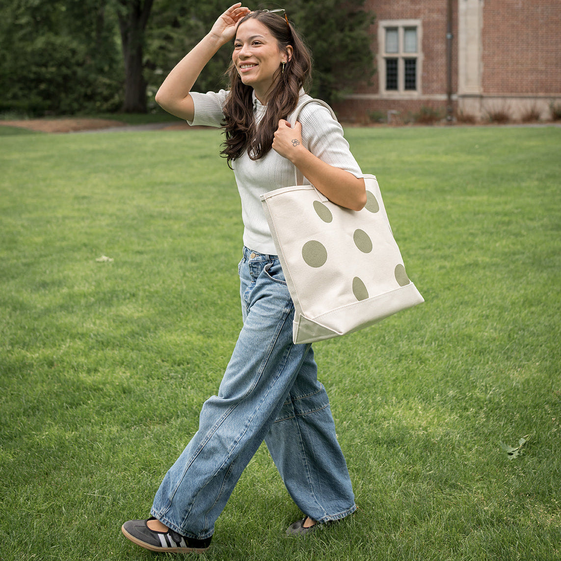 Woman walking on grass holding a polka dot tote bag in front of a brick building.