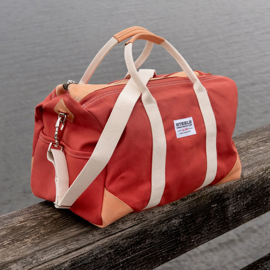 Red duffel bag with beige handles and straps on a wooden surface with a blurred water background