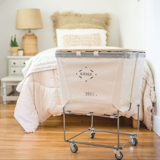 A natural canvas laundry sorter on wheels, placed at the foot of a bed with white bedding.