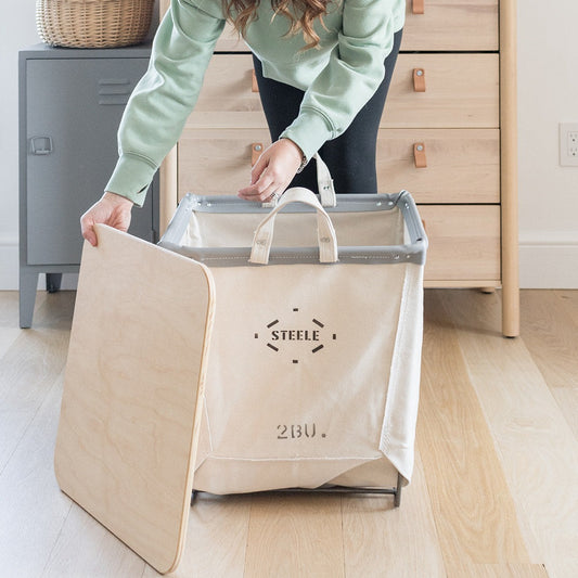 A person is holding a beige square-shaped canvas carry basket with a steel frame on a wooden floor. The basket has a transparent bottom and is labeled with the brand 'Steele' and the size '2 Bu'.