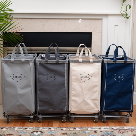 Four laundry baskets with handles in gray, black, beige, and blue, branded 'Steele', on a wooden floor.