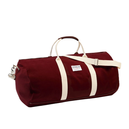 A burgundy canvas duffel bag with cream-colored straps and a white brand label that reads 'STEELE'.