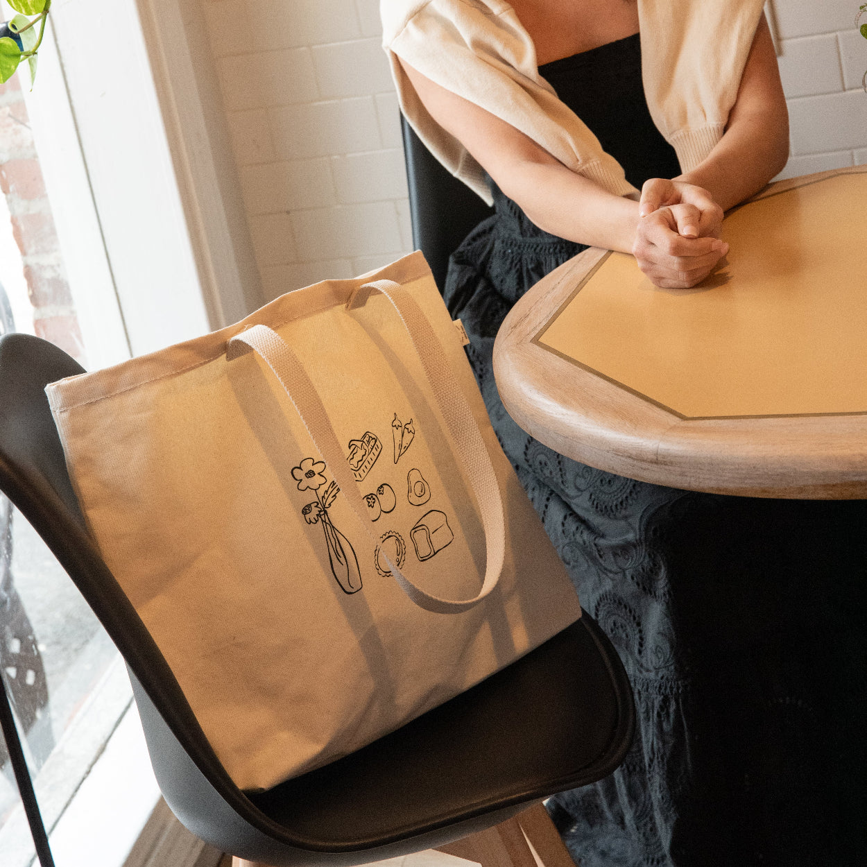 Woman sitting at a table with a beige tote bag featuring illustrations.