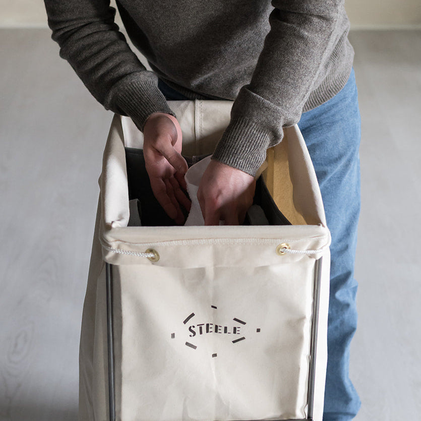 A person in a gray sweater and blue jeans is shown from the waist down, placing or retrieving items from a cream canvas laundry hamper with a metal frame and wheels. The brand name 'STEELE' is printed on the front of the hamper.