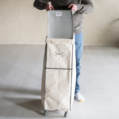 Man holding a large gray bag with a beige rolling cart in a minimalistic room.