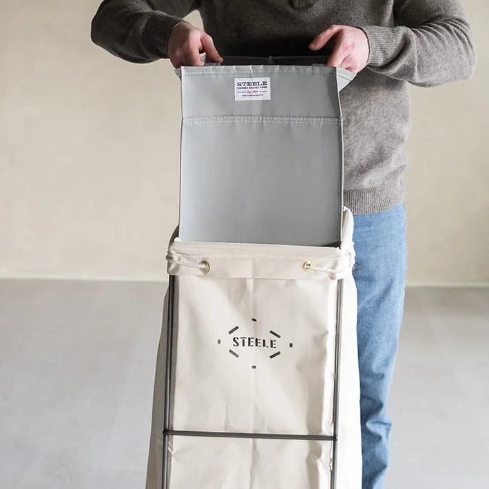 A person wearing a gray sweater and blue jeans stands behind a tall, wheeled fabric storage bag with a gray top panel and a beige lower section. The bag has a black logo reading 'STEELE on the front and is photographed in a minimalist indoor setting with a beige wall and light gray floor.