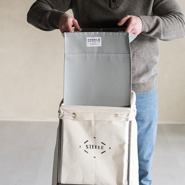 A man wearing a gray sweater and blue jeans stands indoors behind a tall beige fabric cart with wheels, holding the open top of the cart. The cart has a visible logo reading 'STEELE'.