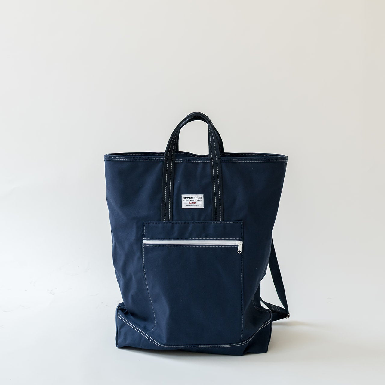 A navy blue canvas tote bag with black handles, a front zippered pocket, and white contrast stitching, photographed against a plain off-white background.
