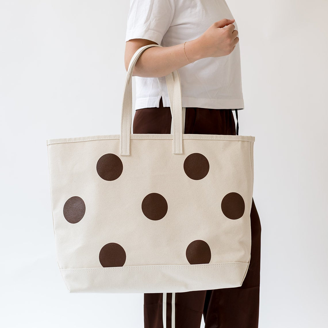 A person wearing a white t-shirt and dark brown pants with white side stripes holds a large cream canvas tote bag decorated with dark brown polka dots, set against a plain white studio background.