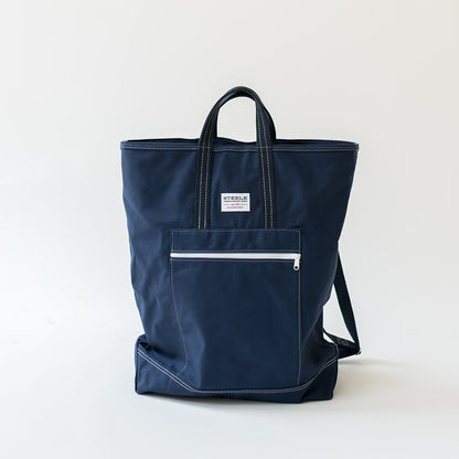 A navy blue canvas tote bag with white contrast stitching, a front zippered pocket, and a small white brand label, photographed on a clean white background.