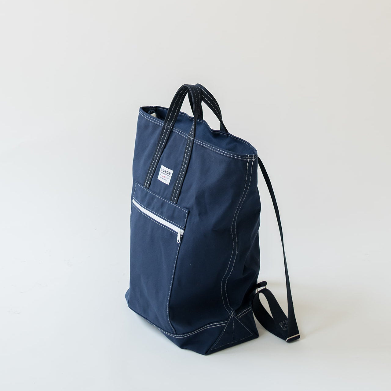 A navy blue canvas tote bag with black straps and white contrast stitching, featuring a front zippered pocket, photographed on a clean off-white background.