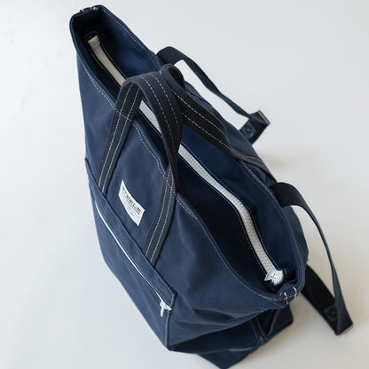 A navy blue canvas tote bag with black straps and white contrast stitching, shown from an overhead angle on a light gray background.