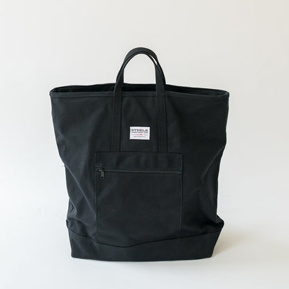 A black canvas tote bag with dual top handles, a front zippered pocket, and a small white brand label, photographed against a clean off-white background.