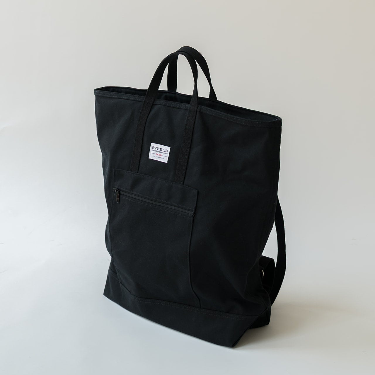 A black canvas tote bag with dual top handles, a front pocket, and a small white brand label, photographed on a clean light gray studio background.