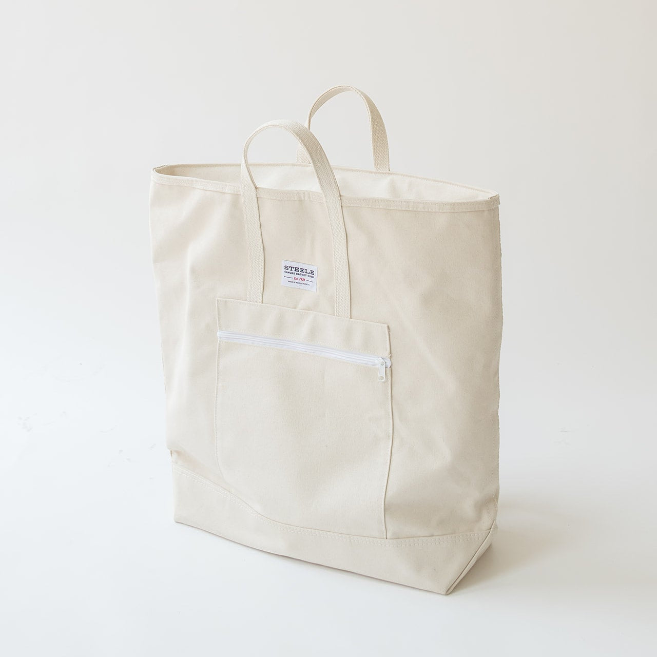 An off-white canvas tote bag with dual top handles, a front zippered pocket, and a small blue brand label, photographed on a clean white background.