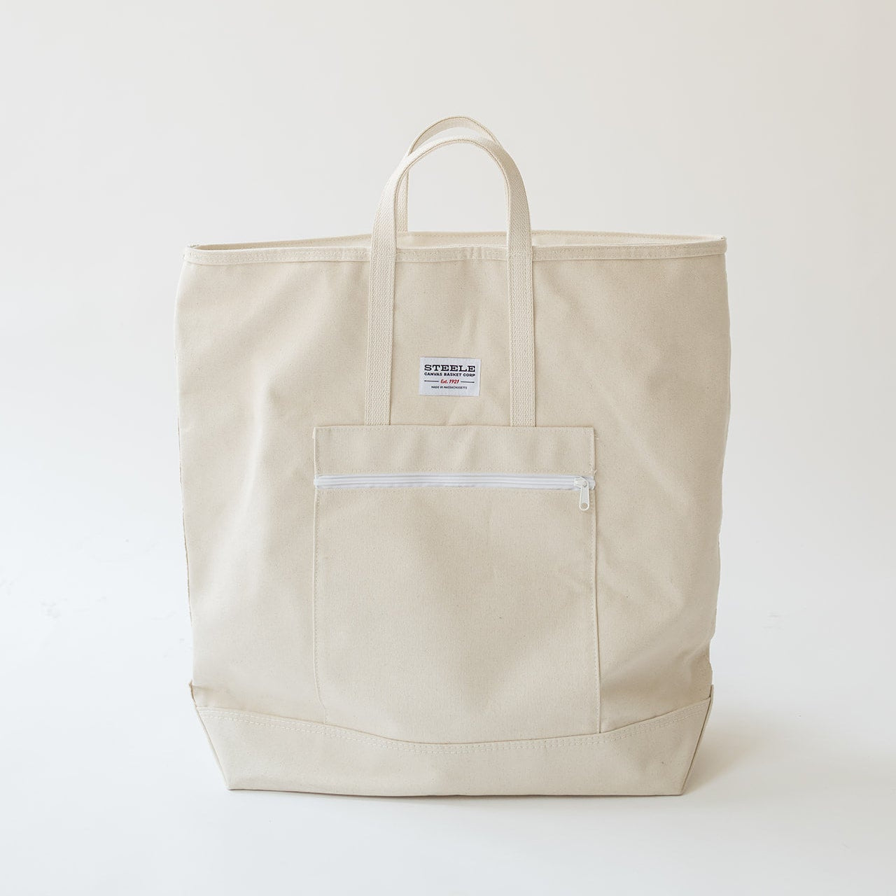 A light beige canvas tote bag with a front zippered pocket and a small white brand label, photographed against a plain off-white background.