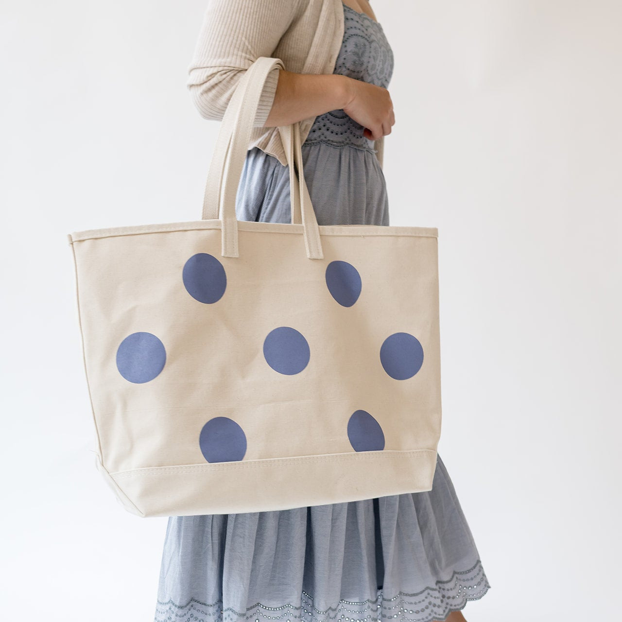 A person wearing a light gray dress holds a large cream canvas tote bag with blue polka dots against a plain white background.
