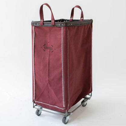 A burgundy fabric laundry hamper with dark gray trim and handles, mounted on a metal rolling cart with gray wheels. The brand name 'STELLE' is printed on the front.