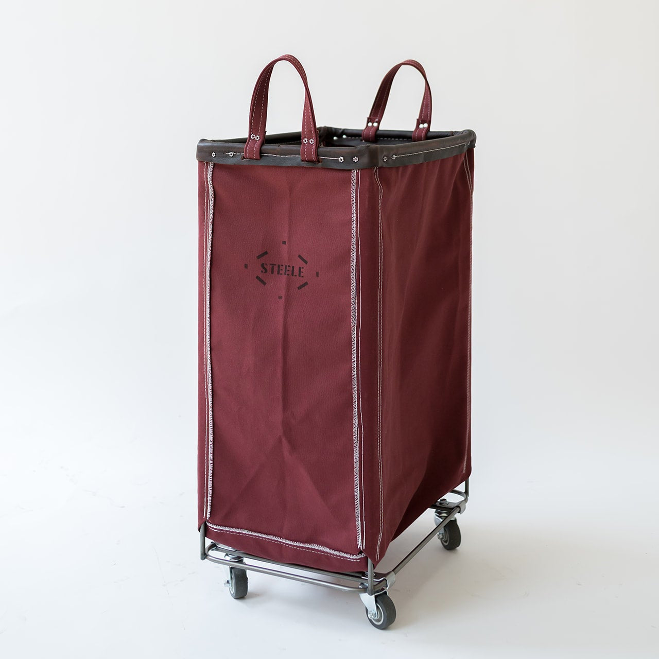 A maroon fabric laundry hamper with dark brown leather handles and trim, mounted on a silver metal rolling frame with casters. The front features a black printed logo that reads 'STEELE'.