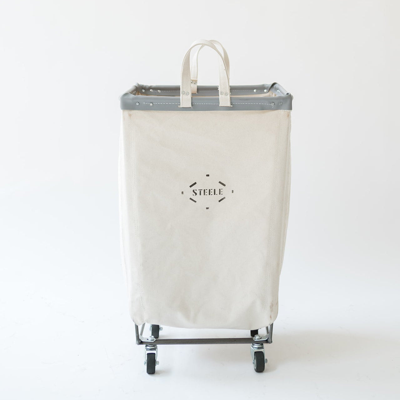 A tall canvas laundry hamper with a gray metal frame and wheels, featuring a top handle and a black 'STEELE' logo printed on the front, set against a plain white background.