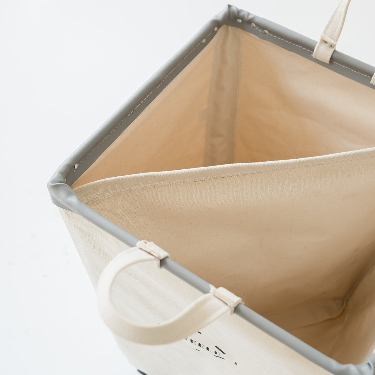 An off-white canvas laundry hamper with gray reinforced edges and cream-colored handles, shown from an elevated angle on a white background.