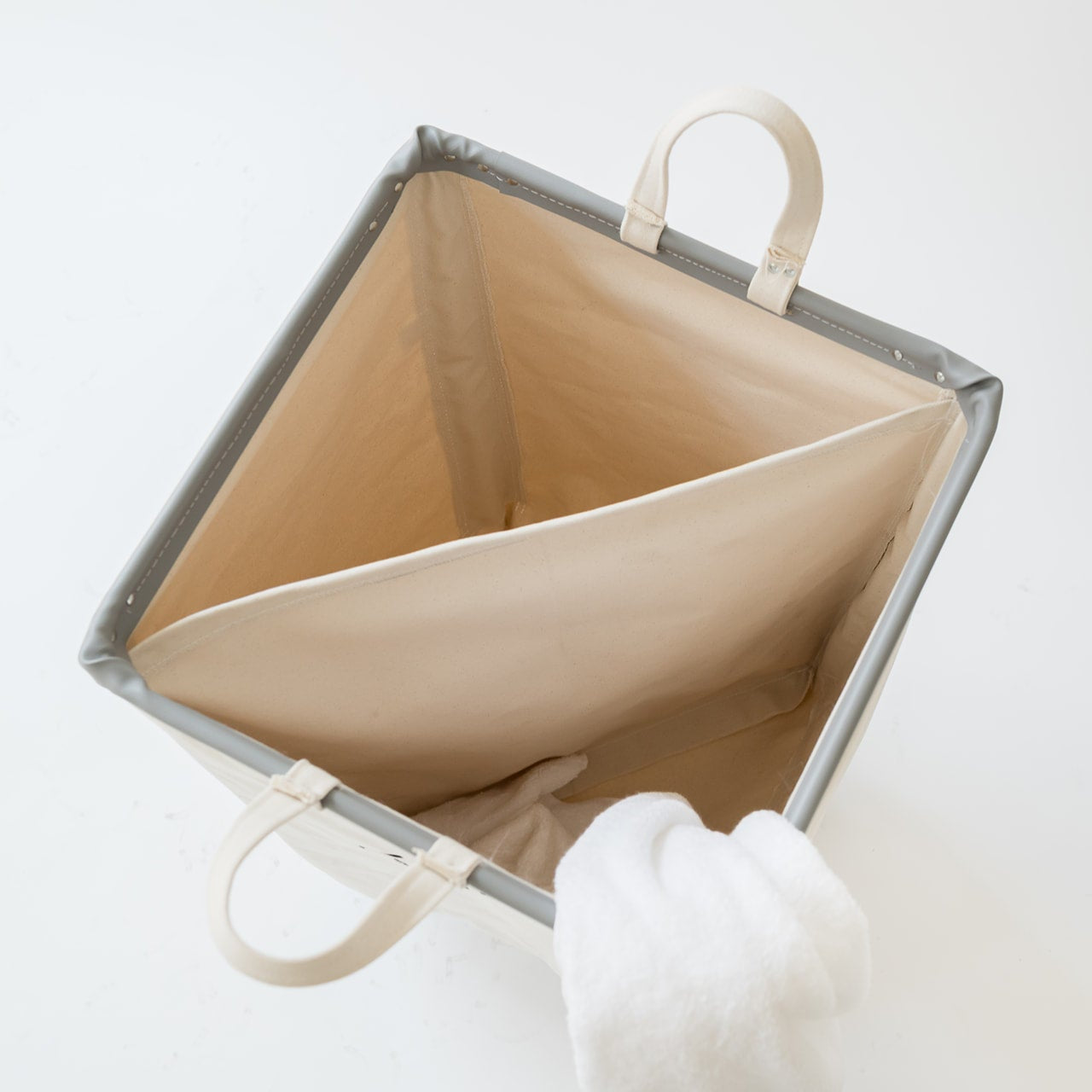 A square fabric storage bin with a gray exterior and beige interior, shown from an overhead angle on a white background. A white textured item is tucked into the bin.