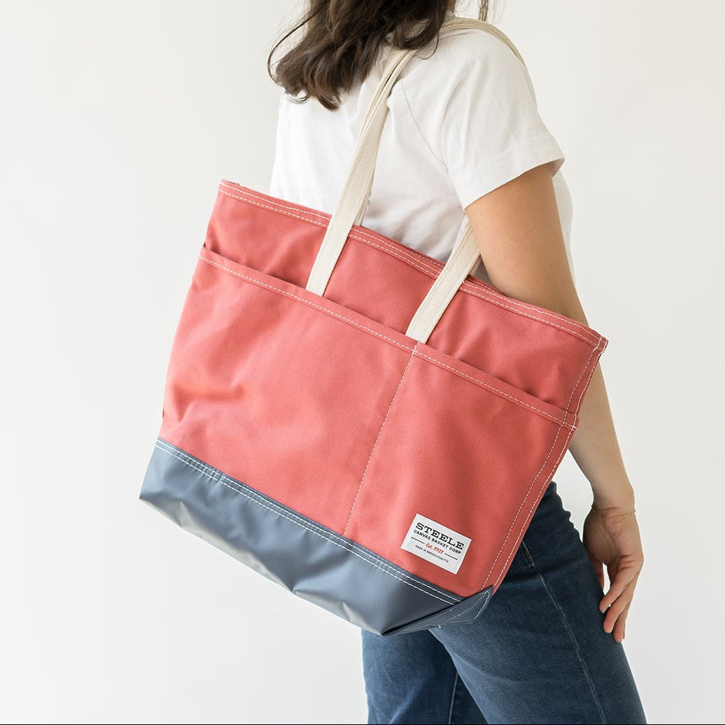 A woman wearing a white t-shirt and blue jeans is shown from behind carrying a large two-tone tote bag. The bag has a coral pink upper section and a slate blue lower section, with cream-colored shoulder straps.