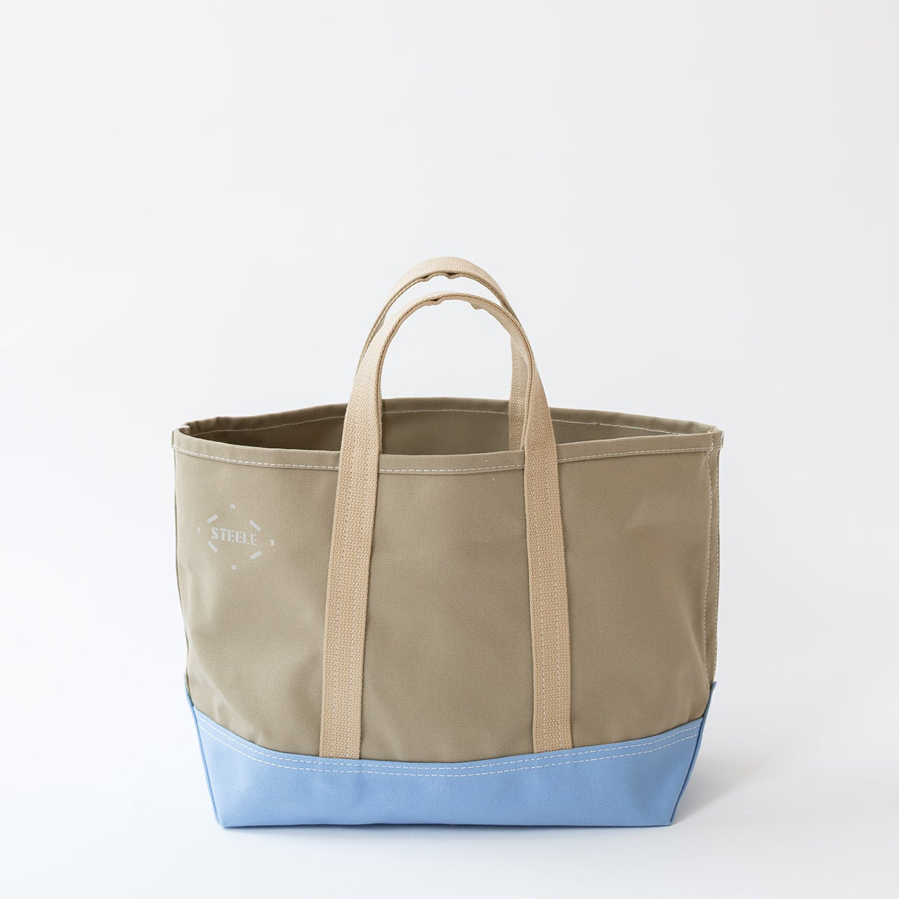 A canvas tote bag with a beige upper body and handles, a light blue bottom base, and a small white embroidered logo on the front, set against a plain white background.