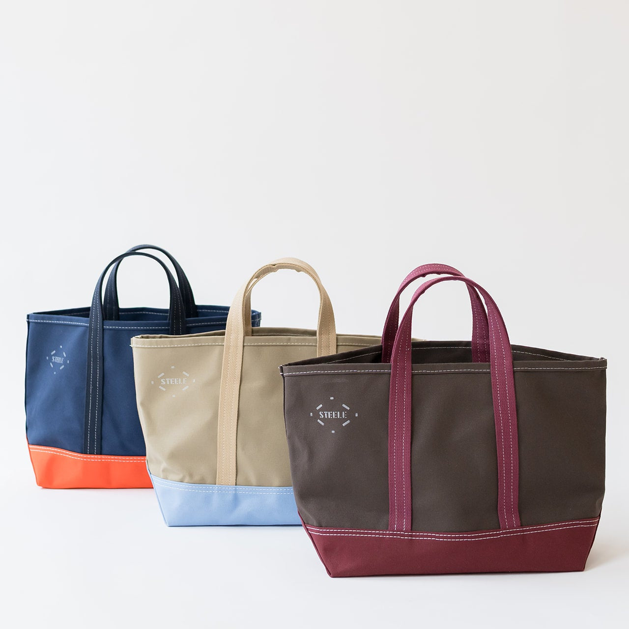 Three canvas tote bags with dual top handles are displayed in a row against a clean white background. The bag in the foreground is dark brown with burgundy handles and a burgundy bottom panel.
