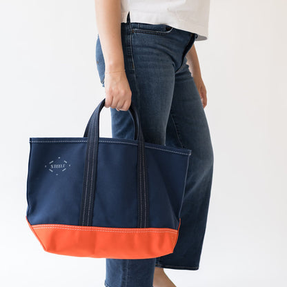 A navy blue tote bag with a bright orange bottom panel and double top handles, held by a person wearing blue jeans and a white t-shirt against a plain white background.