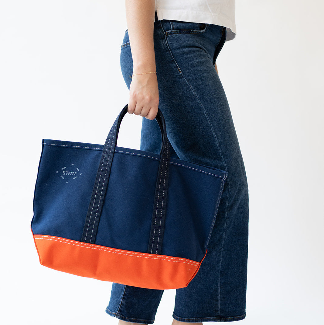 A person wearing a white t-shirt and dark blue jeans holds a navy blue canvas tote bag with a bright orange bottom panel and black handles against a plain white background.