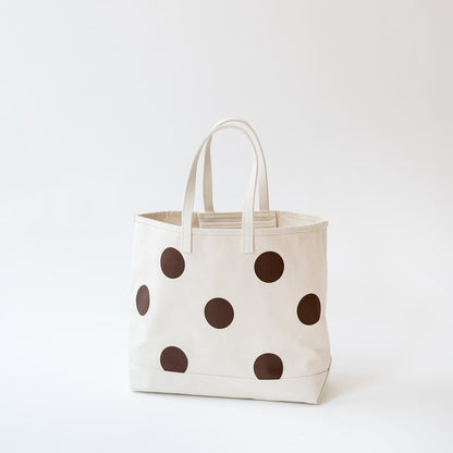 An off-white canvas tote bag with large dark brown polka dots and dual top handles, photographed against a clean white studio background.