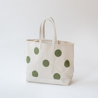 An off-white canvas tote bag with large sage green polka dots and matching fabric handles, photographed against a clean, light gray studio background.