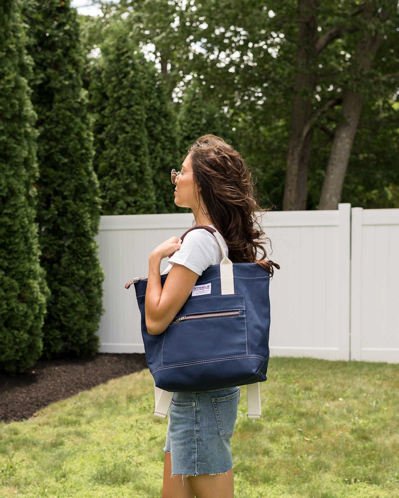 Woman carrying a navy blue tote bag outdoors with greenery in the background