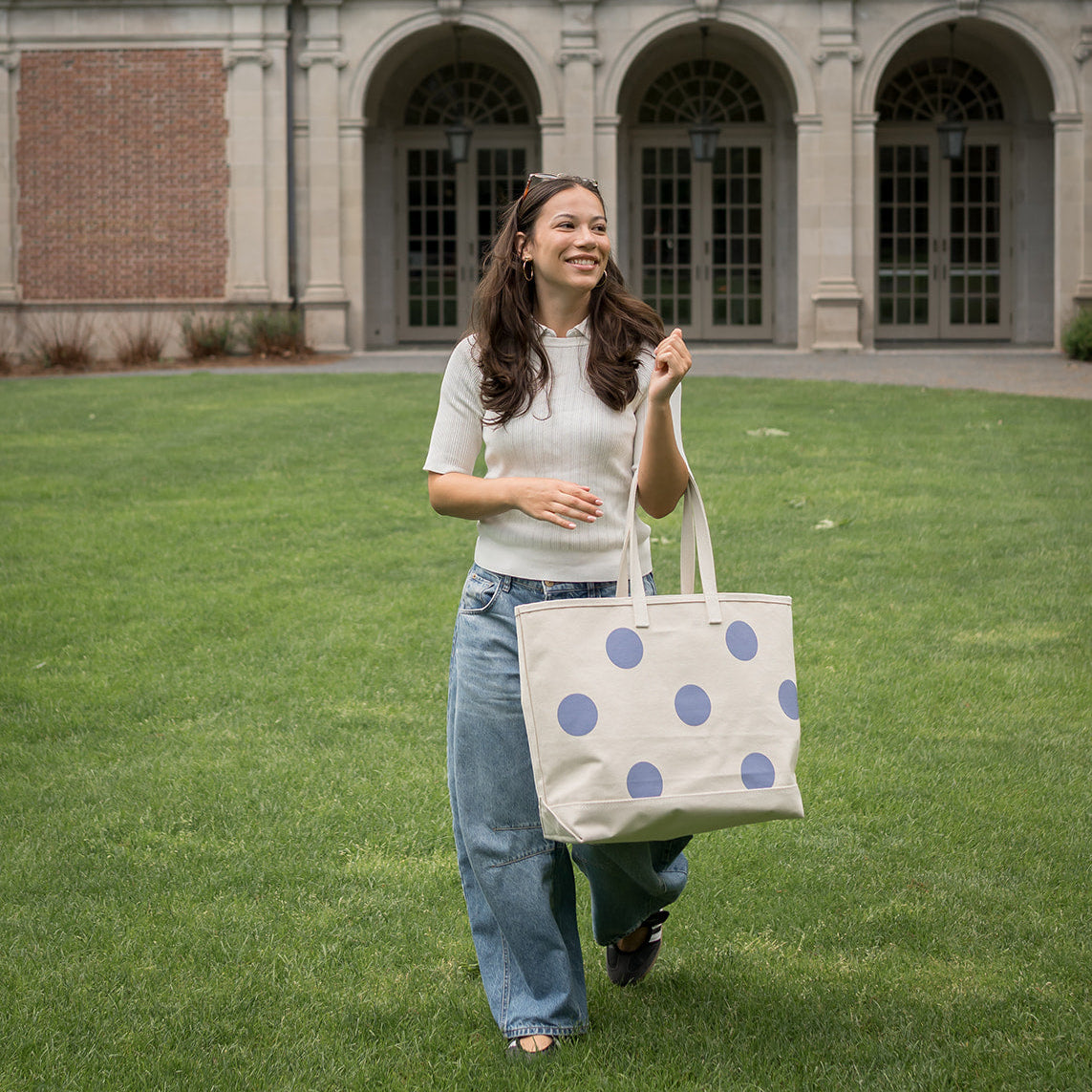 Woman holding a polka dot tote bag on a grassy area in front of a brick building.