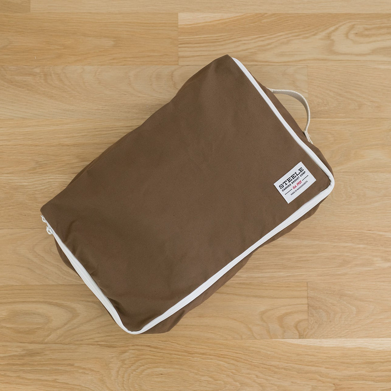 A brown fabric travel luggage tag or packing cube with white piping and a small white brand label, placed on a light wood floor.