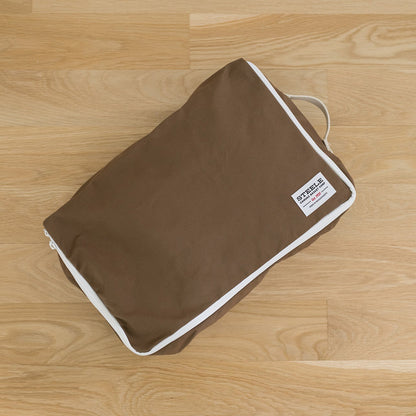 A brown fabric travel luggage tag or packing cube with white piping and a small white brand label, placed on a light wood floor.