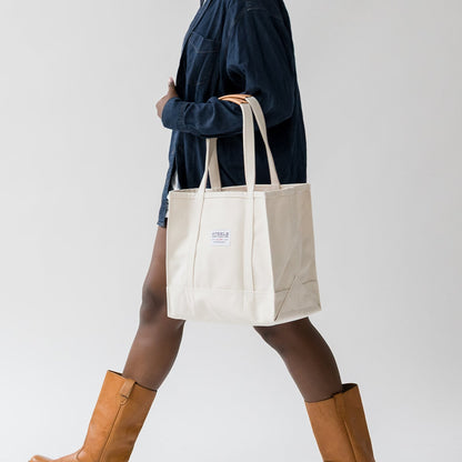 Person holding a white tote bag with a visible brand logo, wearing brown boots on a plain background