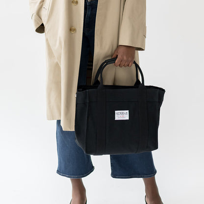 Person holding a black tote bag with a visible brand label on a white background
