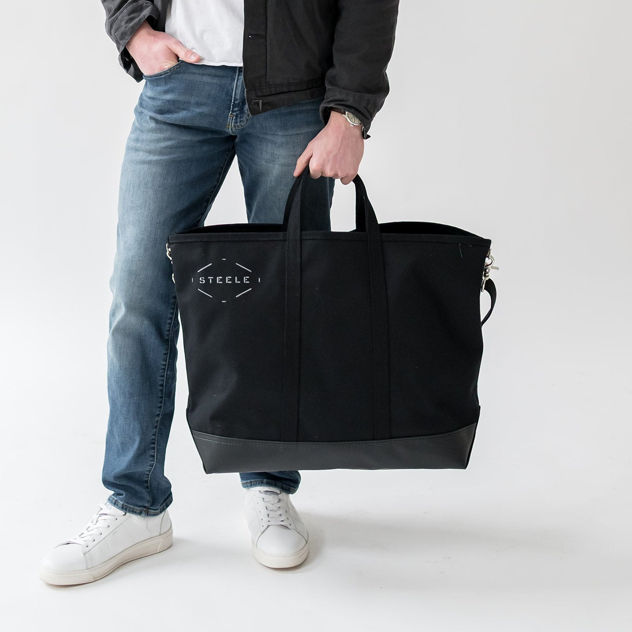 Person holding a black tote bag with 'Steele' branding on a white background