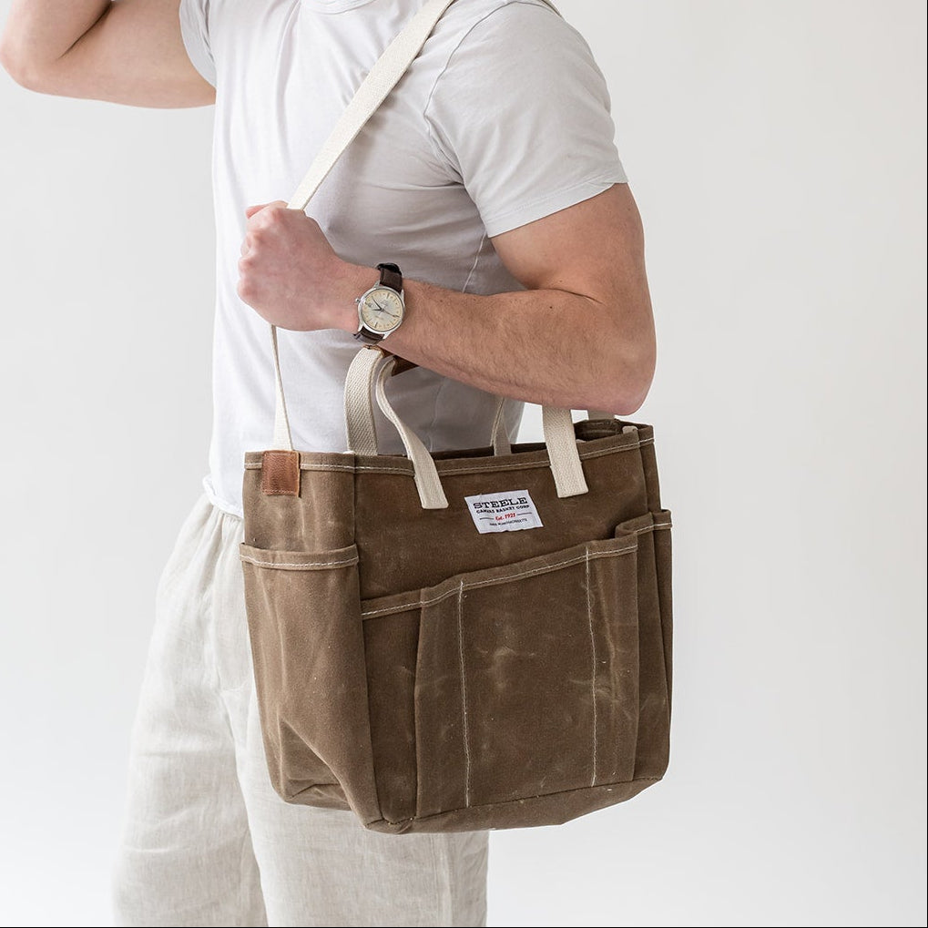 Person wearing a white shirt and light-colored pants with a brown leather bag over their shoulder against a plain background.