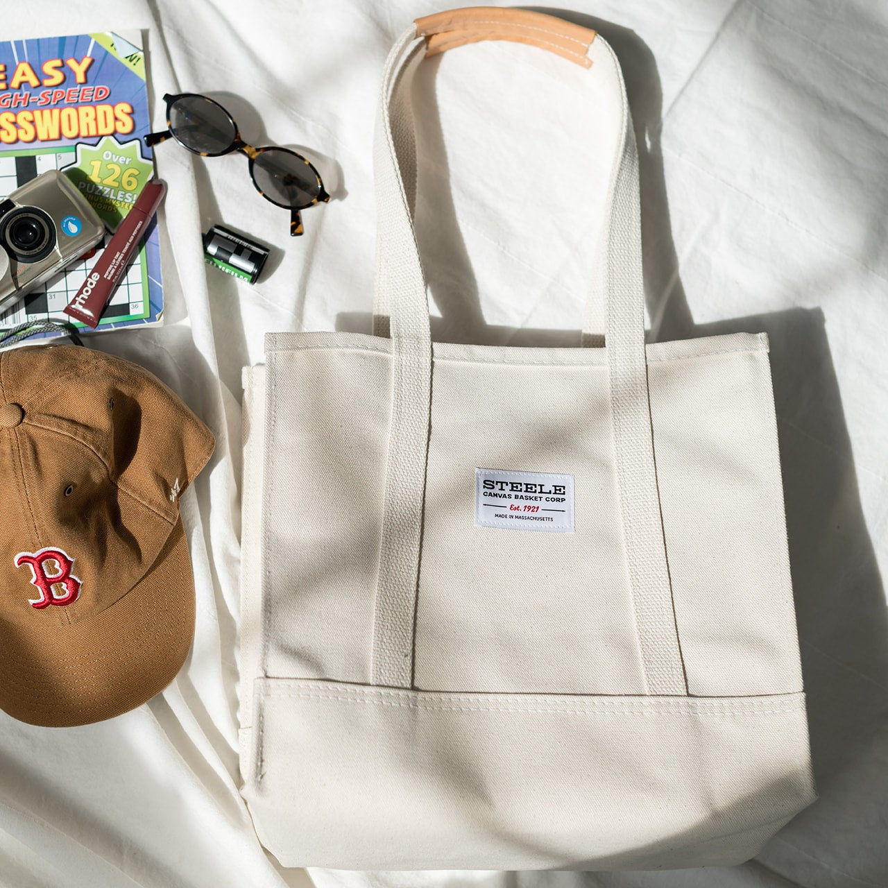 Beige tote bag with a cap, camera, and other items on a white surface
