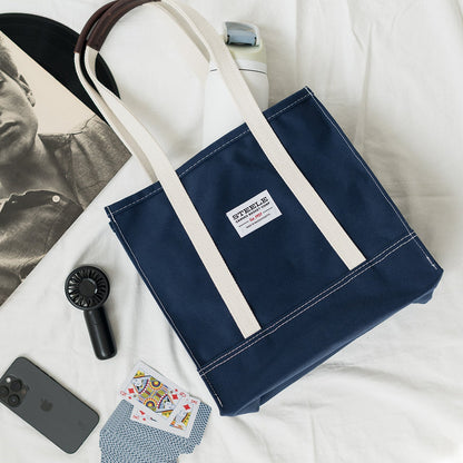 Navy tote bag with white handle on a white surface with a newspaper, phone, sunglasses, and cards.