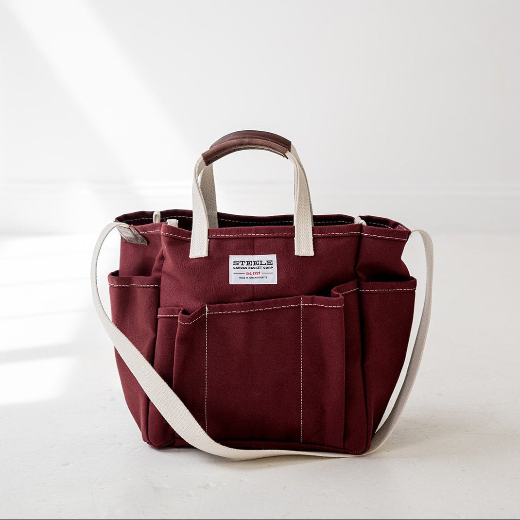 A burgundy canvas utility tote bag with cream-colored handles and shoulder strap, featuring multiple front pockets and a white brand label that reads 'STEELE'. The bag is photographed on a clean white floor against a white wall with soft diagonal sunlight shadows.