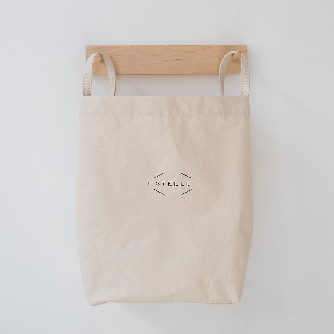 Beige tote bag with 'Steele' logo on a wooden hanger against a white background