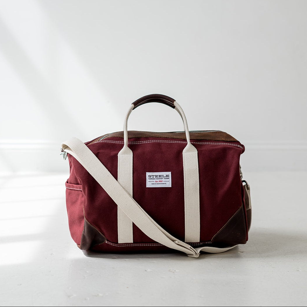 A burgundy and cream canvas duffel bag with a top handle and adjustable shoulder strap, standing on a white floor against a white wall with soft diagonal shadows.