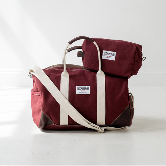 A burgundy canvas duffel bag with cream-colored straps and dark brown leather accents, featuring a matching small zippered pouch. The bags are displayed on a light gray floor against a white wall with soft window shadows.