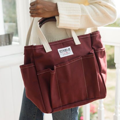 A burgundy canvas utility tote bag with cream handles and a white brand label reading 'STEELE' carried by a person wearing a cream sweater and blue jeans.