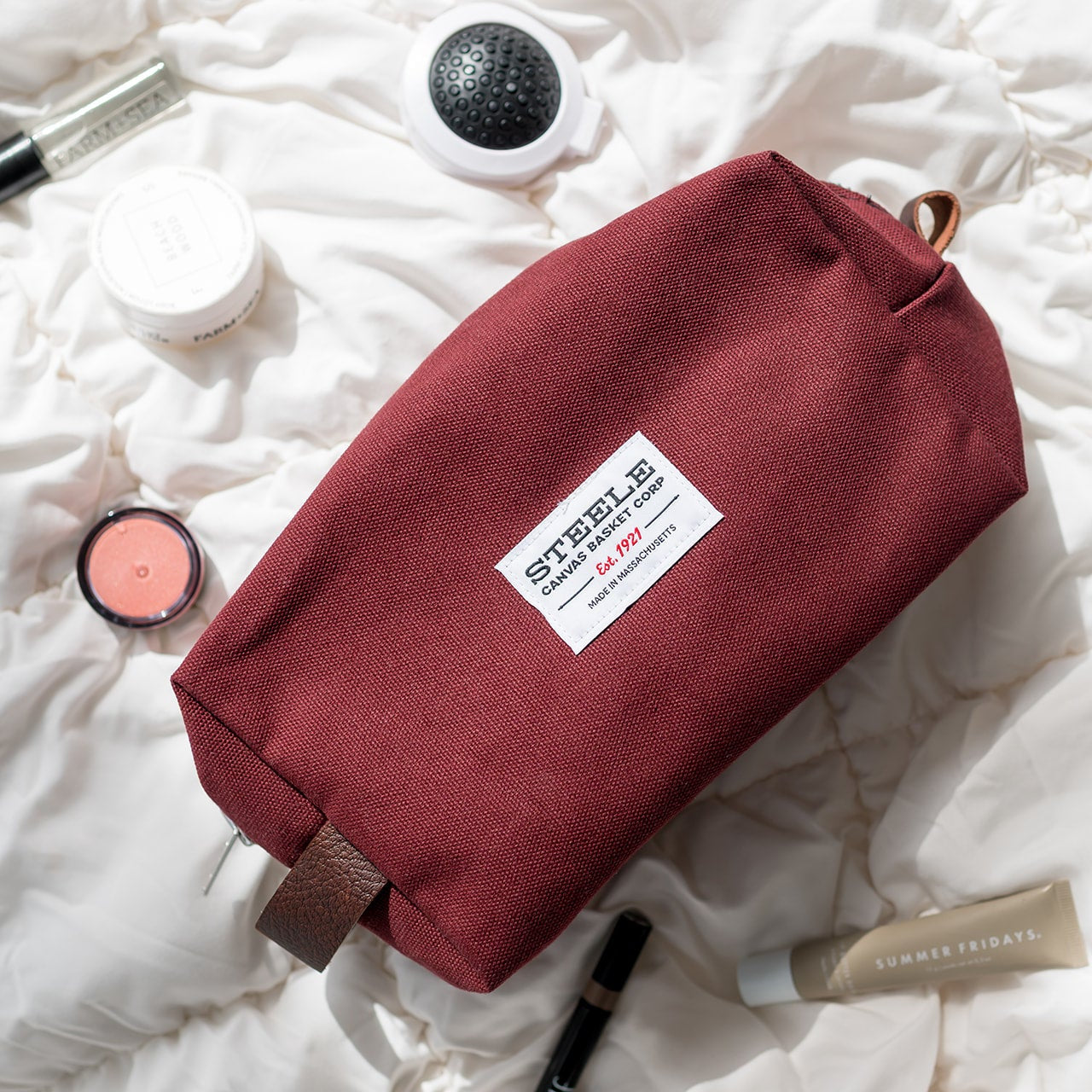 Red toiletry bag with a brand label on a white textured surface with cosmetic items.
